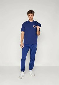 Jaunas vyras stovi su mėlynu marškinėliu ir tokiomis pačiomis mėlynomis Adidas sportinėmis kelnėmis, per petį laiko mėlyną drabužį, matomi balti sportbačiai.