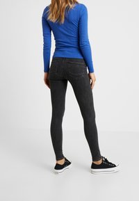 Mujer vestida con una camiseta de manga larga azul, jeans ajustados negros y zapatillas blancas y negras, de pie contra un fondo blanco liso.