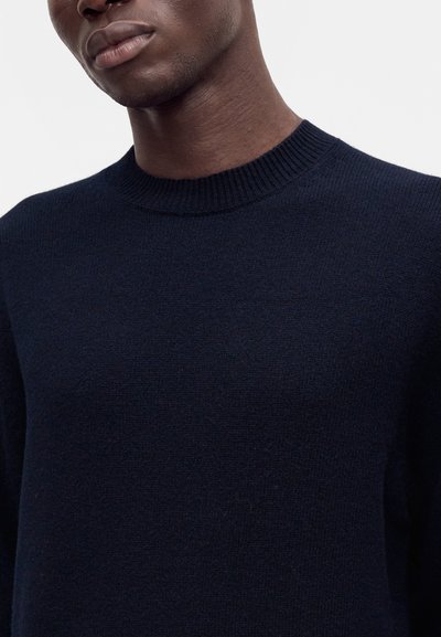 Gros plan d'une personne portant un pull à col rond bleu marine foncé sur un fond clair uni.
