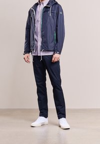 Marineblauwe lichte jas met een capuchon en groene accenten, gecombineerd met een geruit overhemd, marineblauwe broek en witte sneakers. Soepele textuur en casual ontwerp.