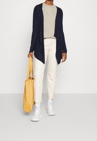 Cardigan blu navy con maniche lunghe e orlo asimmetrico, indossato su una camicia beige, abbinato a pantaloni crema e una borsa a tracolla gialla.