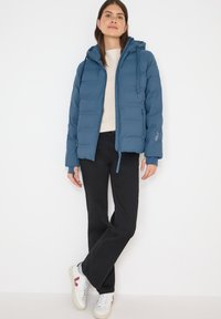 Puffet blå hættejakke med elastiske manchetter, lynlåslukning foran og sidelommer, båret over en beige sweater og sorte bukser. Hvide sneakers inkluderet.