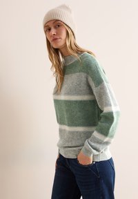 Grön och grå randig tröja med en texturerad stickad mönster, med hög krage och ribbade ärmslut, i kombination med mörkblå jeans.