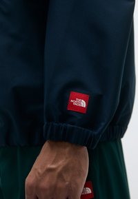 Marineblå jakke med elastiske mansjetter; har et rødt merke med "The North Face"-logoen. Glatt tekstur med en avslappet passform.
