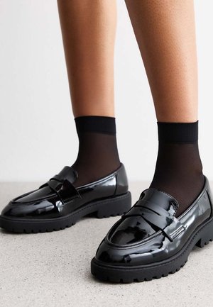 Zwarte gepatineerde leren loafers met een chunky zool, voorzien van een decoratieve riemdetail en gedragen met doorzichtige zwarte sokken.