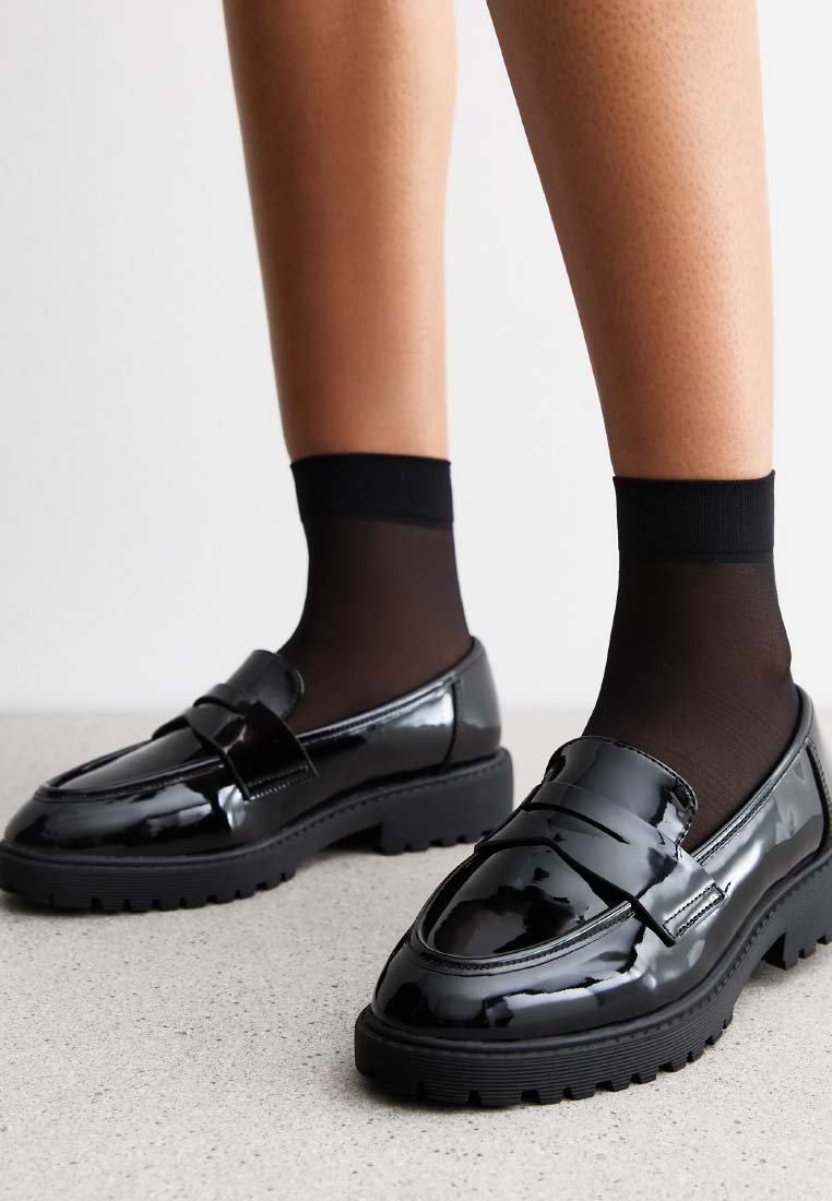 Mocassins en cuir verni noir avec une semelle épaisse, dotés d'un détail de sangle décoratif et portés avec des chaussettes noires transparentes.