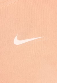 Materiał Nike w kolorze brzoskwiniowym o prążkowanej fakturze, z wyraźnym białym logo Swoosh. Zbliżenie ukazujące miękkość materiału.
