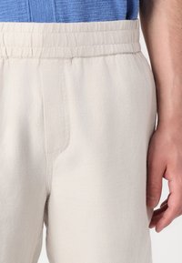 Pantalones de cintura elástica color beige claro, hechos de una tela suave. Presentan bolsillos laterales y una textura lisa, con costuras visibles a lo largo de las costuras.