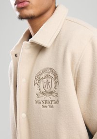 Cream wool-blend jacket with a folded collar, snap buttons, and embroidered logo featuring "Athletic Chains", "Manhattan", and decorative accents.
