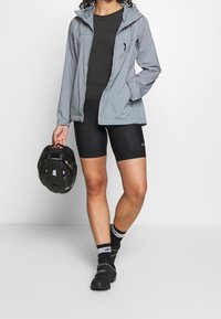 Ljusgrå, lättviktsjacka med huva, bärs över en mörk, ribbad topp och svarta cykelshorts; håller en svart cykelhjälm. Svarta skor och strumpor.