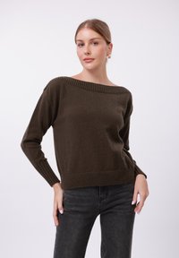 Dunkelbrauner Strickpullover mit weitem, geripptem Halsausschnitt und langen Ä rmeln. Die Textur wirkt weich mit einer lockeren Passform, kombiniert mit schwarzen Jeans.