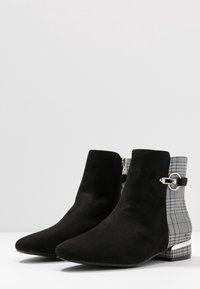 Černé semišové kotníkové boty s špičatou špičkou, které mají panel z kostkované látky a stříbrné kovové akcenty na boku a patě.