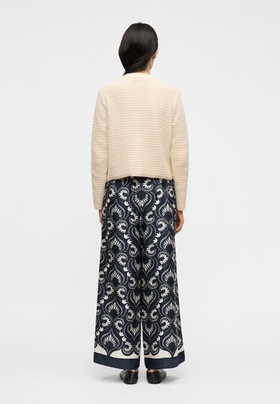 Γυναίκα που φαίνεται από πίσω, φοράει κρεμ πουλόβερ πλεκτό, μπλε σκούρο παντελόνι με λευκά λουλουδένια σχέδια και μαύρα επίπεδα παπούτσια, σε λευκό φόντο.