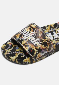 Sandale slide noire avec motif baroque jaune et or, chaînes et logo blanc "Buffalo" sur la bride et la semelle.