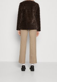 Brun fuskpälsjacka med vertikala ränder, med en avslappnad passform och rund halsringning, tillsammans med beige byxor och svarta klackskor.