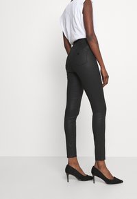 Persona vestida con jeans ajustados negros y tacones altos negros, de pie contra un fondo blanco liso, de espaldas a la cámara.