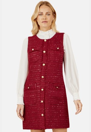 Mujer con vestido sin mangas de tweed rojo con botones dorados y dos bolsillos en el pecho y dos en la cadera, combinado con una blusa blanca de manga larga.