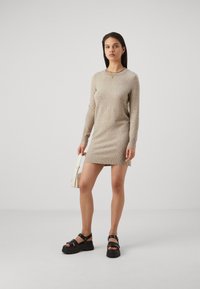 Adīts, bēšs džemperkleitas ar garām piedurknēm un apaļu dekoltē; apvienots ar masīvām melnām sandālēm un mazu baltu rokassomu.