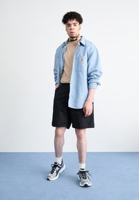 Hellblaues Hemd mit Knöpfen, beiges T-Shirt, schwarze Shorts und graue Sneakers mit Akzenten. Modell steht vor einem schlichten, neutralen Hintergrund.