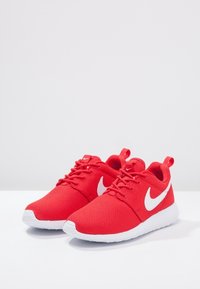 Červené síťované tenisky s bílým symbolem Nike, kulatá špička, šněrovací design a poutko. Mají texturovaný svršek a polstrovanou bílou podešev.