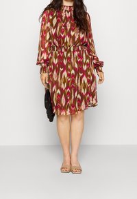 Krátké šaty s květinovým vzorem v odstínech růžové, hnědé a krémové. Dlouhé rukávy s elastickými manžetami a nařaseným pasem. Nosené s černou taškou.