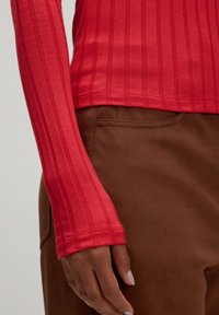 Rotes geripptes Langarmoberteil mit enganliegenden Ärmeln, kombiniert mit braunen weichen Stoffhosen. Das Oberteil hat eine glatte Textur und dezente horizontale Streifen.