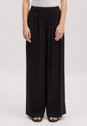 Frau trägt schwarze Hosen mit weitem Bein und elastischem Bund sowie schwarze Sandalen, stehend vor hellem einfarbigem Hintergrund.