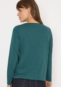 Donna con capelli castani che indossa un top a righe orizzontali turchese e verdi a maniche lunghe e pantaloni scuri, che volge le spalle e guarda di lato.