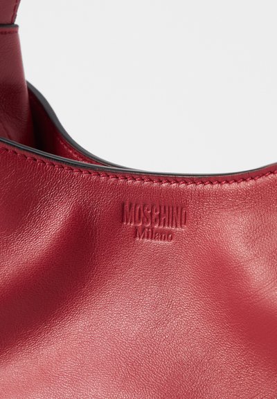 Sarkans ādas roku soma ar gludu, teksturētu virsmu un reljefu "Moschino Milano" logo. Iezīmē kontrastējošas šuves ap malām.