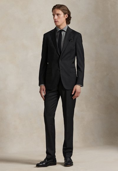 Junger Mann, der einen dunkelgeschnittenen Anzug, schwarze Krawatte, graues Hemd und schwarze elegante Schuhe trägt, vor einem schlichten beigen Hintergrund.