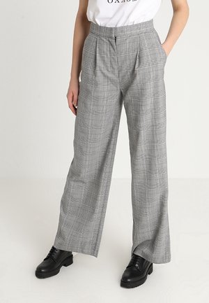 Frau trägt grau karierte Hosen mit weitem Bein und vorderen Falten, kombiniert mit schwarzen Schnürschuhen und einem weiß eingesteckten Hemd.