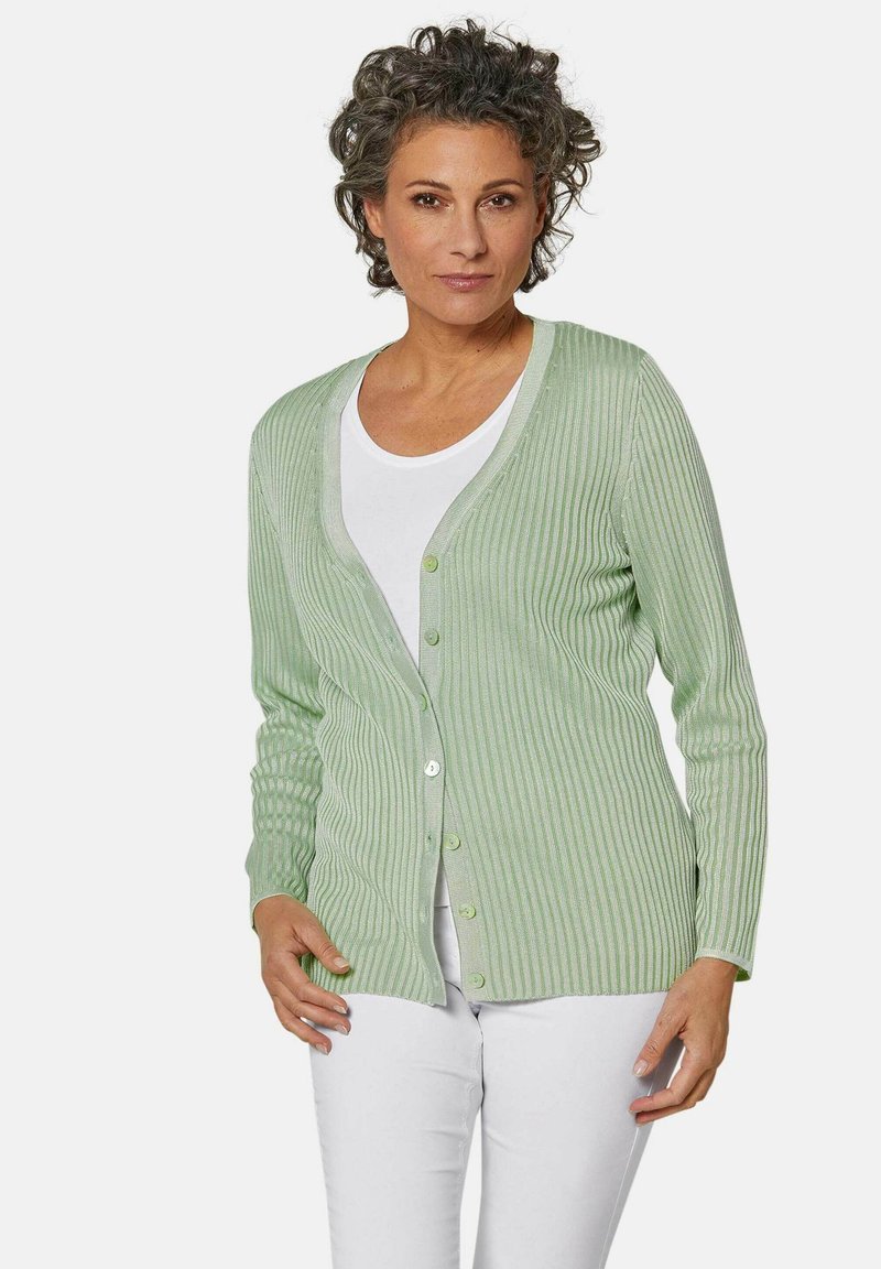 Hellgrüne, gerippte Strickjacke mit V-Ausschnitt, langen Ärmeln und Knöpfen. Getragen über einem weißen Oberteil, kombiniert mit weißen Hosen. Glatte Textur.