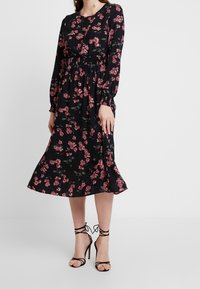 Black floral midi dress with long sleeves, featuring a smocked waist and pink flower patterns. Paired with strappy black high-heeled sandals.