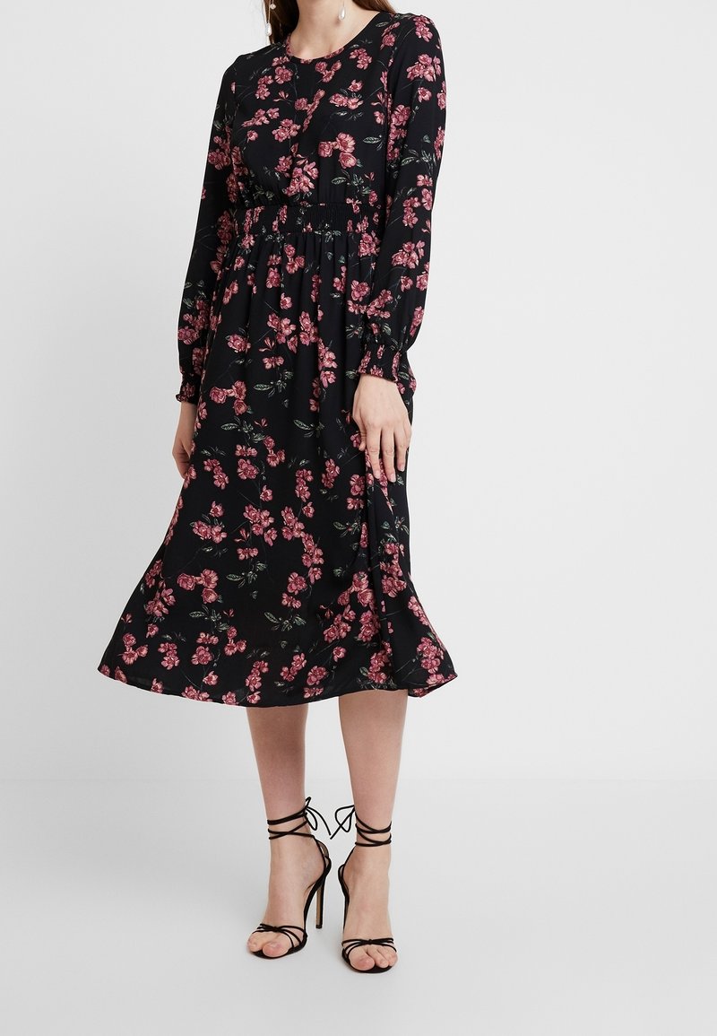 Black floral midi dress with long sleeves, featuring a smocked waist and pink flower patterns. Paired with strappy black high-heeled sandals.