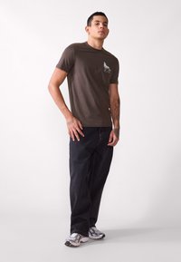 Bruine katoenen t-shirt met een wolf graphic op de borst, gecombineerd met donkere denim broeken en zilveren sneakers, tegen een neutrale achtergrond.