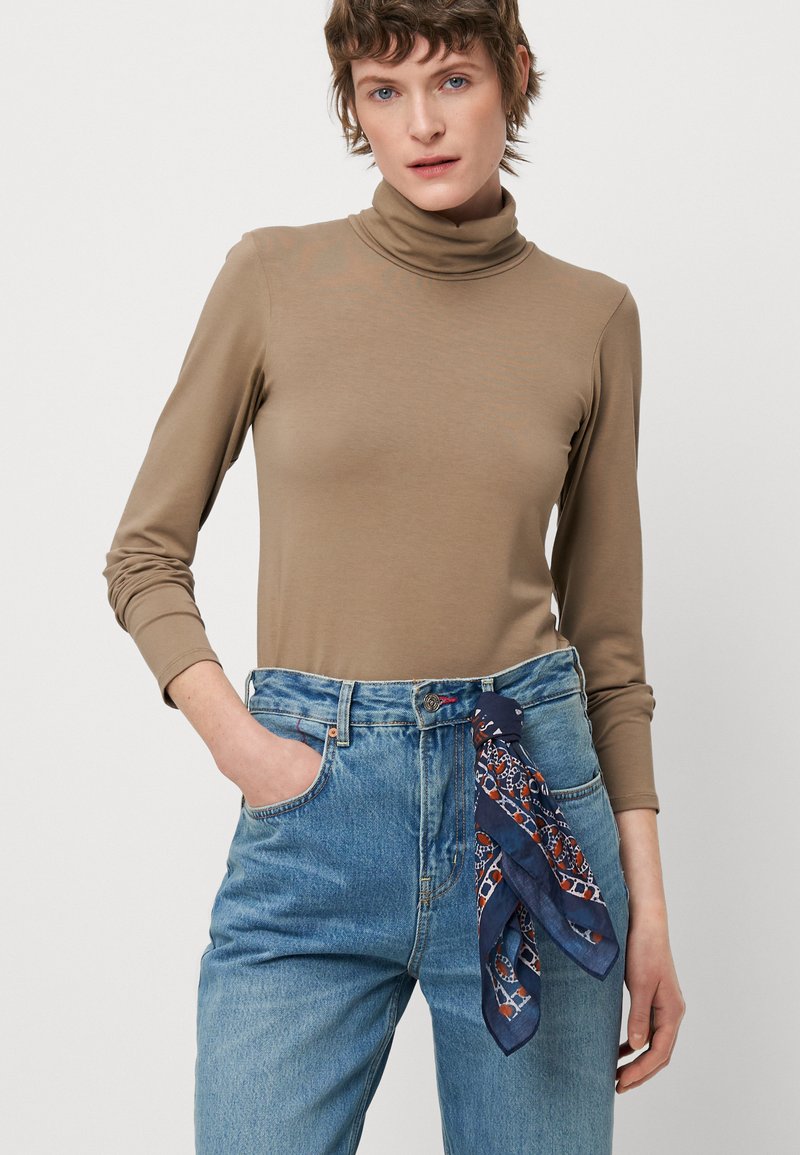 Top de cuello alto en color beige de manga larga combinado con jeans de mezclilla azul de tiro alto. Una bufanda estampada en azul marino realza la cintura. Textura suave.