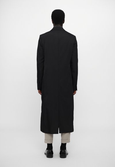 Caban negru lung, cu o siluetă dreaptă, textură netedă și design minimalist. Gulerul are un detaliu ușor plisat, purtat peste pantaloni deschiși.