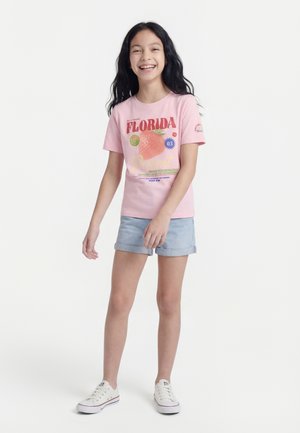 Χαμογελαστή κοπέλα με μακριά σκούρα μαλλιά, φορώντας ροζ μπλουζάκι με γραφικά της Φλόριντα, ανοιχτόχρωμα τζιν σορτς με γυρισμένα μπατζάκια και λευκά αθλητικά παπούτσια, στέκεται σε λευκό φόντο.