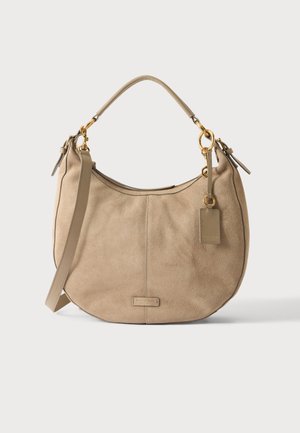 Beige suede shoulder bag with curved shape, gold hardware rings, a short handle, a long strap, and a small leather tag attached.