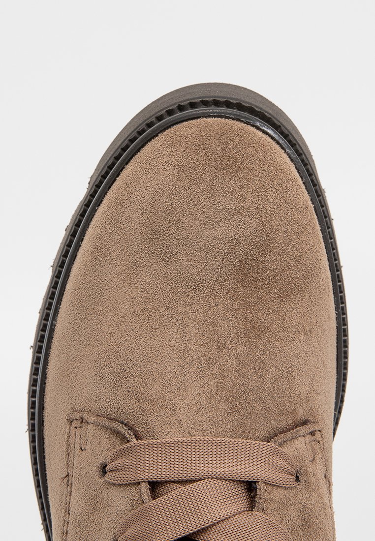 Bruine suède schoen met een ronde neus en dikke zwarte zool, met bijpassende bruin veters die in een strik zijn gestrikt, gezien vanuit een top-down perspectief.