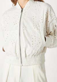 Kvinna som bär en vit broderad bomberjacka med hålmönster, frontdragkedja, elastiska ärmslut och matchande vita byxor.