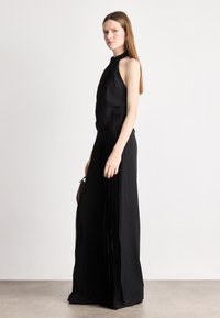 Μαύρο φόρεμα με ψηλή λαιμόκοψη και πλισέ κορσέ που καταλήγει σε ρέουσα φούστα. Μαλακό ύφασμα με λεία υφή. Το μοντέλο κρατά μια μικρή, σκούρα τσάντα.