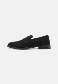 Mocassins en daim noir avec une texture lisse, un bout rond et une lanière décorative. Ils présentent un talon bas et solide ainsi que des détails de couture subtils.