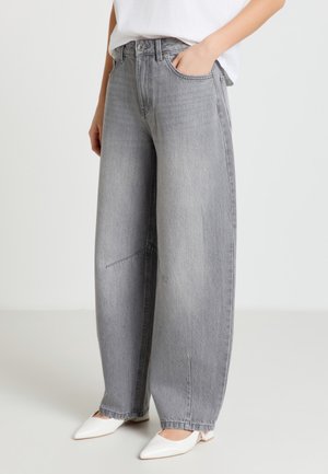 Personne portant un jean gris clair ample et des chaussures blanches pointues à talons bas, une main dans la poche, sur un fond uni.