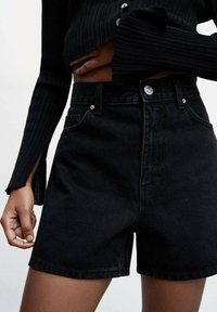 Černé denimové šortky s vysokým pasem mají klasický střih, přední kapsy a kovové zapínání. Hladká textura s minimálním zdobením.