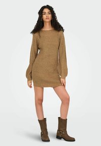Robe en tricot couleur marron clair, avec de longues manches bouffantes, un col rond et une silhouette ajustée. Associée à des bottes marron jusqu'aux genoux.