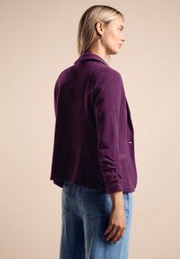 Lila kavaj i mjukt tyg, med krage, lös passform och långa ärmar, buren över ljusblå vida jeans, sedd bakifrån.