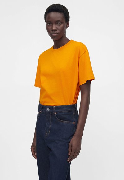 Personne portant un t-shirt à manches courtes orange vif rentré dans un jean taille haute bleu foncé, sur un fond clair uni.