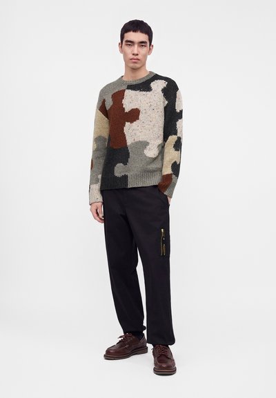 Jeune homme portant un pull multicolore avec des pièces de puzzle, un pantalon noir avec une poche latérale zippée et des chaussures marron à lacets, debout devant un fond uni.