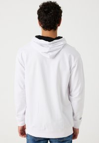 Sudadera blanca con capucha hecha de un tejido suave, que presenta un bolsillo tipo canguro en la parte frontal y puños acanalados, con un forro interior negro en capas visible.